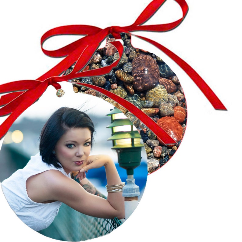 May include: A round Christmas ornament with a red ribbon. The ornament features a photo of a woman with short black hair, wearing a white top, leaning on a railing. The background is a blurry image of a street scene with a streetlight.