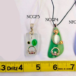 May include: Five glass pendants with different designs and colors, including green, blue, and white. Each pendant has a silver Celtic knot charm and is hanging from a black cord. The pendants are labeled with the text "NCGP1", "NCGP3", "NCCP4", and "NFGP5".