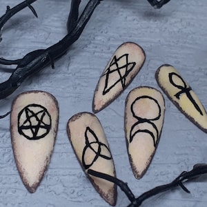Puede incluir: Cinco uñas postizas en forma de almendra con base beige y símbolos negros. Los símbolos incluyen un pentagrama, una estrella dentro de un diamante, un triquetra, medias lunas y un ankh. Ramas oscuras enmarcan las uñas.