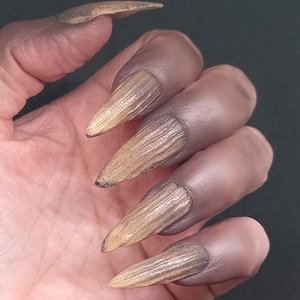 May include: A set of long, pointed artificial fingernails featuring a textured, wood-grain appearance in shades of light brown and tan. The nails have a tapered, claw-like shape with a matte finish, displaying vertical striations that mimic natural wood.