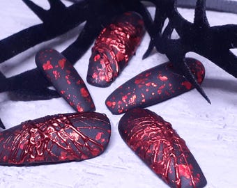 May include: Close-up of five black nail tips with red accents. The nails have a matte finish and are decorated with red foil and a textured, dripping design. The background includes black, branch-like decorations.