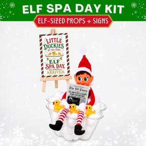 May include: An elf spa day kit featuring a sign on a wooden easel that reads Little Duckies By My Side Elf Spa Day Has Officially Arrived. The kit includes a clear plastic bowl filled with white pom-poms, three yellow rubber ducks, and a newspaper.