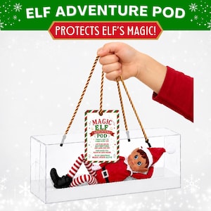 May include: A clear rectangular plastic carrying case with brown rope handles containing a small elf doll wearing a red and white outfit. A rectangular tag hangs from the handles with the text MAGIC ELF ADVENTURE POD and additional text.