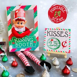 May include: A Christmas elf doll dressed in red and white stripes is peeking out of a red, green, and white kissing booth sign. The elf is holding a sign that says "Kissing Booth". There is a sign that says "Kisses 25¢" and a box of Hershey's Kisses.