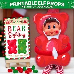 Puede incluir: Un disfraz de oso de goma rojo con una cara de elfo y un letrero que dice "BEAR Sighting". El letrero está sobre un pequeño caballete de madera, con ositos de goma rojos y verdes esparcidos. El texto "PRINTABLE ELF PROPS INSTANTLY DOWNLOAD" está en la parte superior.