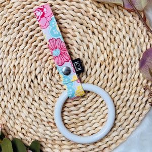 Peut inclure: Un anneau de dentition blanc avec une sangle à motifs floraux. La sangle présente des fleurs roses et jaunes sur un fond bleu clair. L'anneau est fixé avec un bouton-pression argenté. Le fond est une surface tissée brun clair.