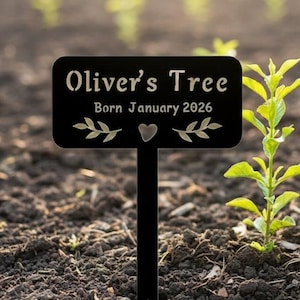 May include: A black metal garden stake with the text "Oliver's Tree Born January 2026" and decorative leaf and heart accents. A small green plant grows beside the stake in the dark soil of a garden.