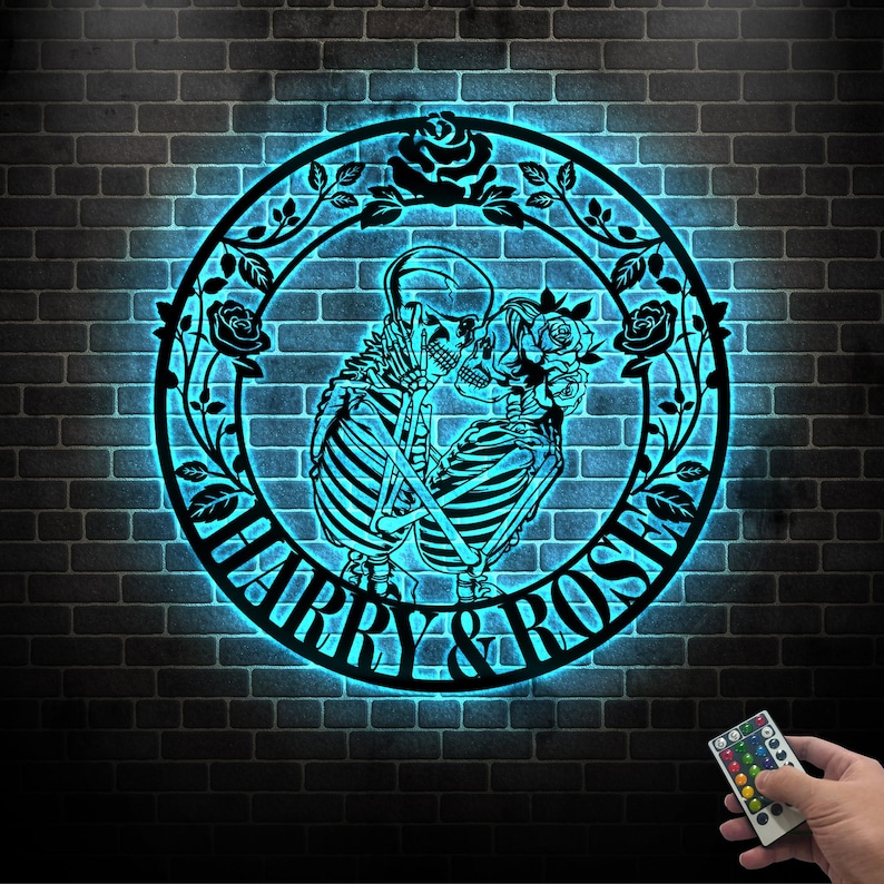 May include: A black metal wall art piece featuring two skeletons embracing within a circular frame. The frame is adorned with roses and leaves, and the words "HARRY & ROSE" are displayed. The art is illuminated with a blue glow against a brick wall.