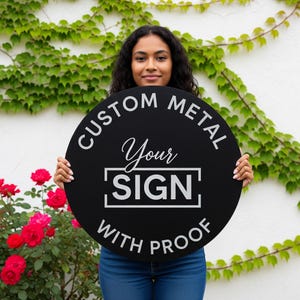 May include: A round, black metal sign with white text that reads "CUSTOM METAL Your SIGN WITH PROOF." The sign is held by a person in front of a white wall covered in green vines and red roses.