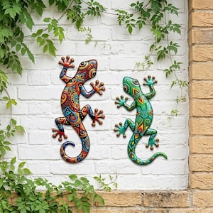 May include: Two metal lizard wall art pieces. One lizard is primarily red, orange, and blue with intricate patterns. The other is green, blue, and yellow. Both are mounted on a white brick wall with green foliage.