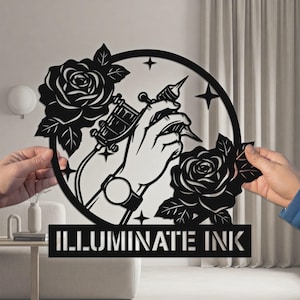 May include: Black metal wall art featuring a hand holding a tattoo machine, surrounded by roses and stars. The text "ILLUMINATE INK" is displayed at the bottom. The design is held by two hands.