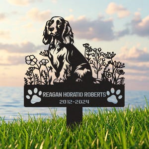 Könnte beinhalten: Schwarze Metallsilhouette eines Spaniel-Hundes, der in einem Blumenfeld sitzt. Das Schild trägt die Aufschrift "Reagan Horatio Roberts 2012-2024" mit Pfotenabdrücken auf beiden Seiten.