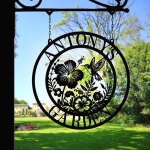 May include: Black metal garden sign with the text "ANTON'S GARDEN" in a circle. The sign features a hummingbird and floral designs.