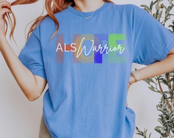 May include: A periwinkle blue short-sleeve t-shirt featuring the text "ALS" in white block letters and "Warrior" in white cursive script. Behind the text, the word "HOPE" is printed in large, colorful, sunburst-patterned letters.