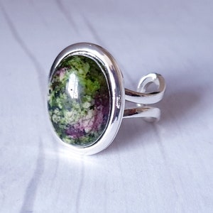 May include: A silver ring featuring a green and pink oval gemstone set in a bezel setting. The ring has a double band.