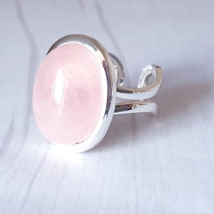 May include: A silver adjustable ring with a large, oval rose quartz gemstone. The pale pink stone is set in a polished silver band with a split shank design. The ring is shown on a light-coloured surface.