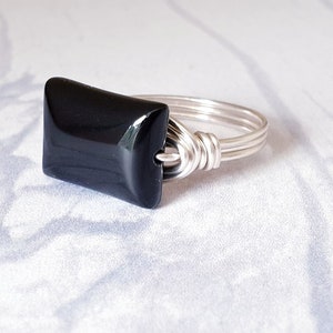 May include: A ring featuring a smooth, rectangular black stone set horizontally. The band is constructed from multiple strands of silver-colored wire that wrap around the stone to secure it in place, creating a layered and textured metal finish.