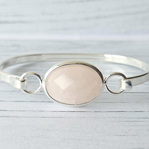 May include: A silver bangle bracelet featuring a central oval-shaped, faceted pale pink stone. The stone is set in a smooth silver bezel and connected to the polished silver band by two circular silver rings on either side of the setting.