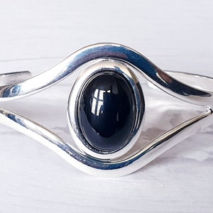 May include: A silver cuff bracelet with a large, oval, black gemstone center. The bracelet has a modern, open design with a polished finish. The gemstone is approximately 1 inch in length.