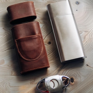 Può includere: Un assortimento di accessori per sigari, tra cui un astuccio per sigari in pelle marrone con tasca, un coperchio abbinato, un tubo per sigari in metallo argentato e un tagliasigari argentato. Gli oggetti sono disposti su una superficie di legno chiaro.