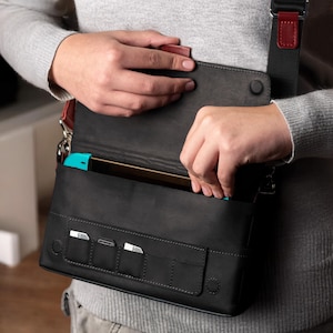 Pode incluir: Bolsa transversal de couro preta com uma alça vermelha e preta. A bolsa tem um fecho de aba e vários bolsos pequenos na frente. Uma pessoa é mostrada abrindo a bolsa para revelar um console de jogos turquesa.