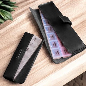 May include: Black leather pill case with a weekly pill organizer inside. The case has a snap closure and a small pocket on the outside. The pill organizer has compartments for each day of the week.