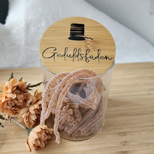 Könnte beinhalten: Ein Glasgefäß mit einem Holzdeckel. Der Deckel hat eine schwarze Silhouette einer Garnrolle und den Text "Geduldfaden". Das Gefäß ist mit braunen, zuckerhaltigen Süßigkeiten gefüllt.