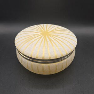 May include: A round, frosted glass trinket box featuring a radiating sunburst pattern in gold. The lid and base are decorated with these gold lines, and a thin, dark metallic band separates the lid from the base, creating a distinct visual contrast.