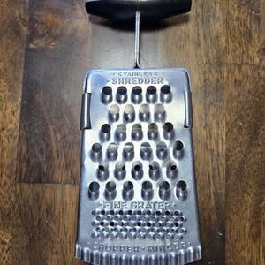 May include: Stainless steel kitchen grater with a black handle. The grater has a rectangular shape with multiple holes for shredding and grating. The text "SHREDDER", "FINE GRATER", and "CHOPPER MINCER" are visible.