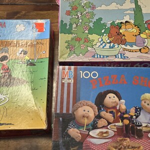 Puede incluir: Tres rompecabezas vintage. Uno presenta una caricatura de Peanuts con el texto "HA HA HA HA HA". Otro muestra a Garfield y sus amigos en una barbacoa. El tercer rompecabezas representa a Cabbage Patch Kids en una pizzería.