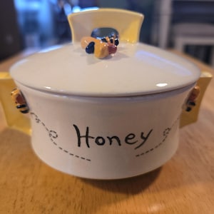 May include: Cream-colored ceramic honey pot with a lid and two handles. The lid is topped with a bee figurine. The word "Honey" is written on the front in black script, with dotted lines and bee graphics.