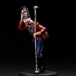 Könnte beinhalten: Eine bemalte Figur eines Soldaten in rot-blauer Uniform, einem schwarzen Hut und einem silbernen Stab. Die Figur steht auf einem schwarzen Sockel.