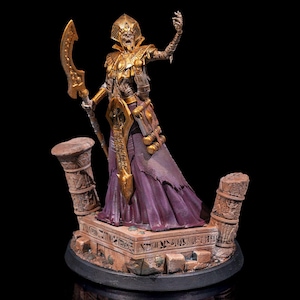 Puede incluir: Una figura miniatura pintada en oro y morado de una figura esquelética que lleva un tocado dorado y sostiene un bastón dorado. La figura está de pie sobre una base de piedra con jeroglíficos.