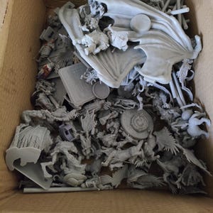 Może przedstawiać: Pudełko wypełnione niepomalowanymi szarymi plastikowymi figurkami, w tym smokami, rycerzami i innymi fantastycznymi stworzeniami. Figurki są gotowe do złożenia i pomalowania.