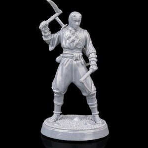 Puede incluir: Figura gris de un ninja con vestimenta tradicional, sosteniendo un arma con cuchilla y una espada. El ninja está de pie sobre una base circular con una superficie texturizada. El fondo es negro.