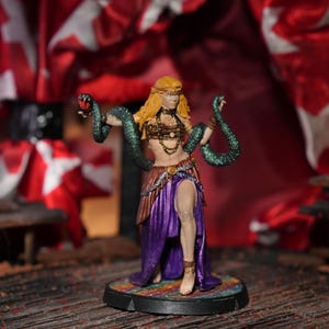 Könnte beinhalten: Miniaturfigur einer Frau mit langen blonden Haaren, geschmückt mit Schmuck und einem lila Rock. Zwei grüne Schlangen sind um ihre Arme gewickelt. Die Figur steht auf einem runden Sockel mit farbenfrohem Design, vor rotem und weißem Hintergrund.