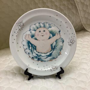 Puede incluir: Un plato de cerámica blanco con un diseño de col azul y un bebé dentro. El plato está decorado con conejos blancos y pequeñas estrellas.