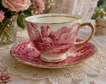 Può includere: Un set di tazza da tè e piattino in porcellana bianca con un dettagliato motivo a trasferimento rosso raffigurante paesaggi rurali con figure e cottage. Il bordo della tazza, il bordo del piattino e il manico sono rifiniti in oro.