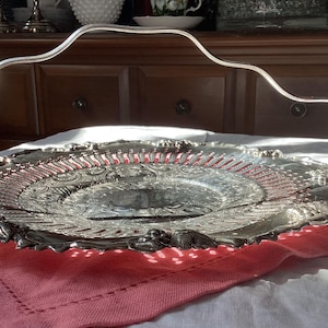 Puede incluir: Una bandeja de servir plateada con un diseño decorativo y un asa de alambre plateada. La bandeja está sobre un mantel rosa.