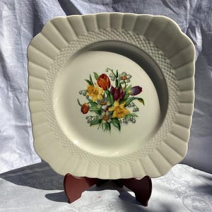 May include: Cream-colored square plate with a floral bouquet design. The plate features a scalloped edge and a textured border. The bouquet includes red, yellow, and purple flowers. A decorative piece for serving or display.