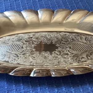 May include: A silver-coloured, oval serving tray with a decorative, scalloped edge. The centre of the tray features an ornate, engraved floral design. The tray is approximately 46 cm long.