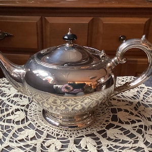 May include: A silver teapot with an ornate design and a black knob on the lid. The teapot is sitting on a white crocheted doily.
