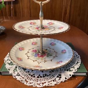 May include: Two-tiered serving tray with floral design. The plates are white with gold trim and a central floral pattern in pink, blue, and yellow. The handle is gold-coloured. The tray is sitting on a white lace doily.