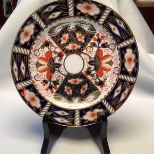 Può includere: Un piatto in ceramica rotondo con centro bianco e un motivo complesso nei toni del blu scuro, arancione e oro. Il design presenta motivi floreali stilizzati, forme geometriche e volute intricate, rifinito con un bordo color oro.