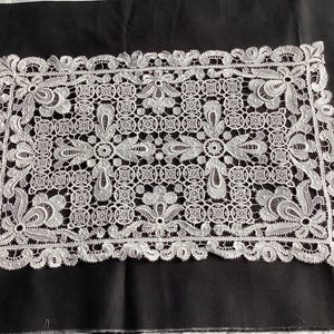 May include: A white lace doily with a detailed floral pattern, rectangular in shape. The doily has a scalloped edge and is displayed on a black surface. The lace design incorporates flowers and geometric elements.