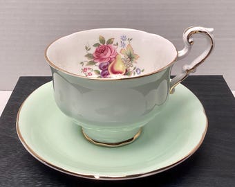 May include: A mint green teacup and saucer set. The teacup features a floral and fruit design with a pink rose and gold trim. The saucer has a matching gold trim. The handle of the teacup is white with gold accents.