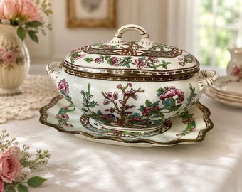 May include: A white ceramic tureen with a matching oval platter, featuring a colorful floral pattern in pink, green, and blue, accented with brown geometric borders, two side handles, and a lid with a central handle for serving food.
