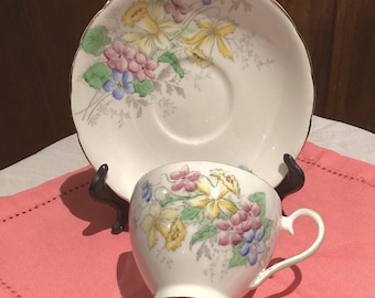 May include: A white teacup and saucer with a floral pattern featuring yellow, pink, and blue flowers. The floral pattern is on a white background with a gold trim around the edge of the saucer.