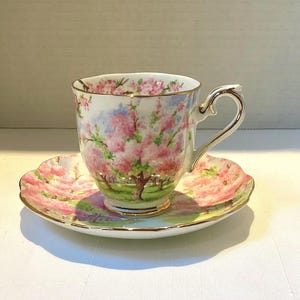 May include: A porcelain teacup and saucer set. The cup and saucer feature a pink floral design with gold trim. The cup has a curved handle and the saucer has a scalloped edge. The set is on a white surface.