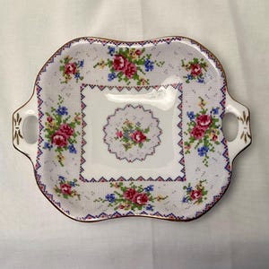 May include: Square, white ceramic serving tray with a floral cross-stitch design. The tray features pink and blue flowers, gold trim, and handles on either side. The tray is approximately 9 inches square.